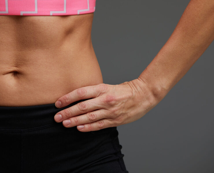 Croped close up body of fit woman wearing shorts and sport top showing slim beautiful stomach and abs in diet. Fitness and healthy lifestyle concept isolated on grey background
