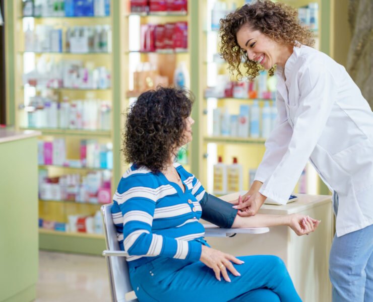 Smiling young female pharmacist in white robe, while standing near sitting lady customer with legs crossed and looking at each other during blood pressure checking near blurred shelves in pharmacy