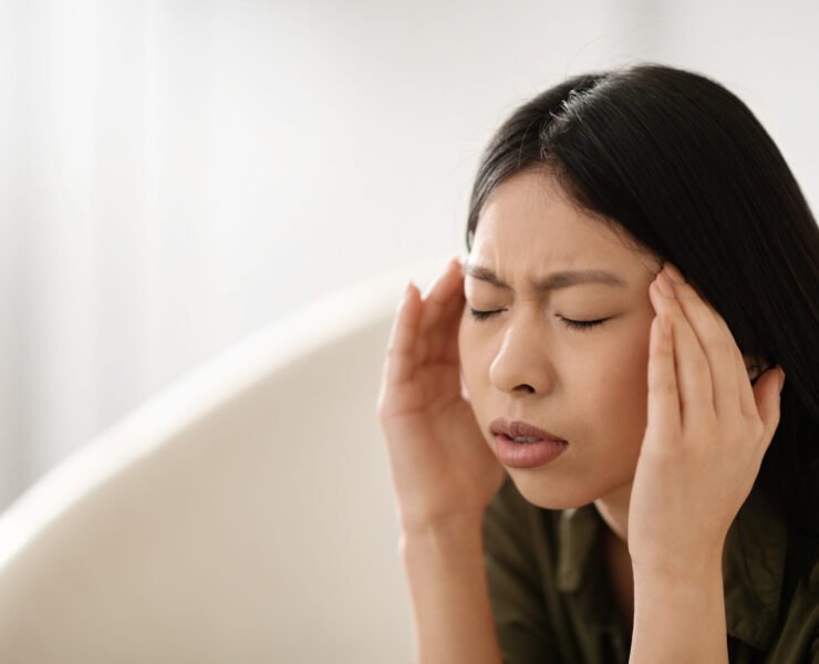 Sick unhappy young asian woman sitting on couch with closed eyes, touching her head, home interior, closeup, copy space, home interior. Migraine, headache, unhealthy lifestyle concept